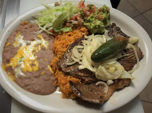 A plate of sliced grilled steak under onions and a fried jalapeno, with rice, beans, pico de gallo, lettuce and guacamole