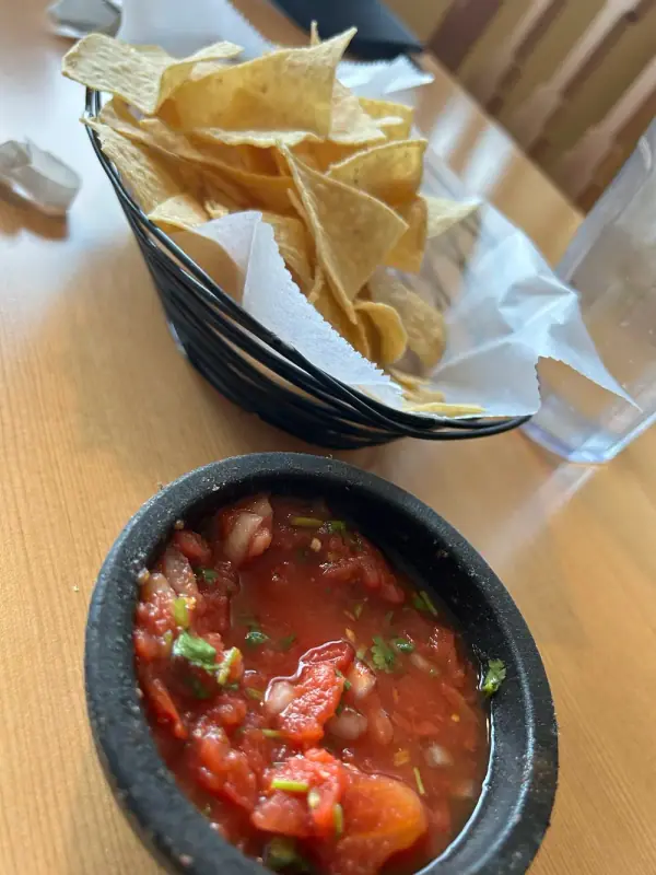 A wire basket of tortilla chips and a black stone bowl of chunky fresh salsa on a wood table