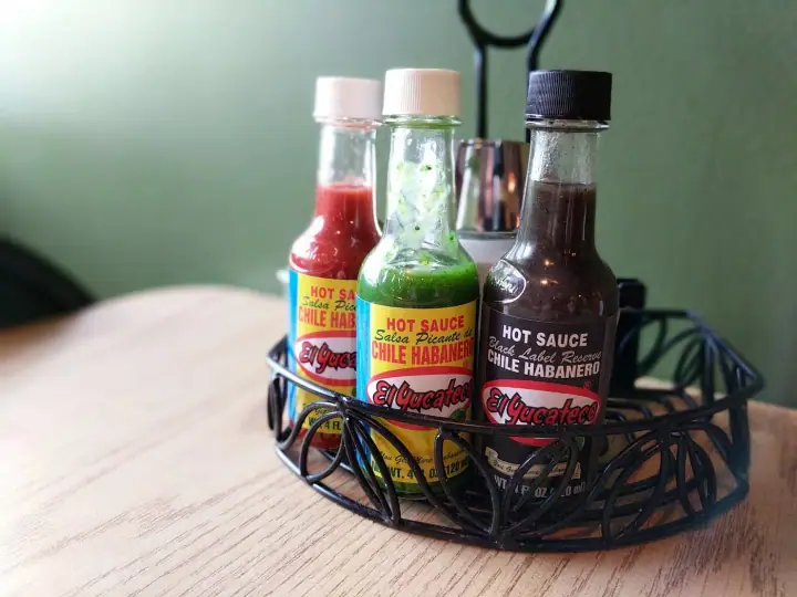 A black wire caddy holding bottles of hot sauce on a wood table in front of a green wall